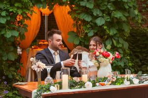 A wedding party celebrates at a decorated head table with floral arrangements and orange drapery in the background.