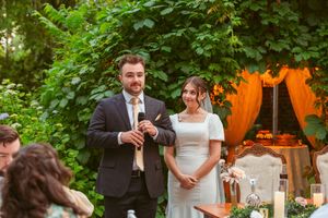Romantic outdoor wedding celebration scene with couple at reception featuring orange decor and greenery backdrop in garden.