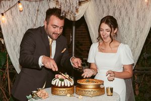 A couple in formal wedding attire cuts into a tiered cake together at an outdoor reception with string lights and white drapes.