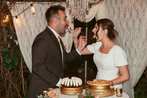 A happy couple shares cake together at their wedding reception in front of a rustic lace backdrop with string lights.