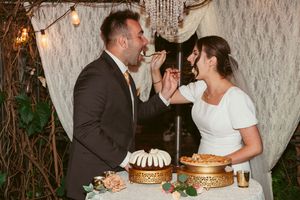 Couple playfully sharing cake at their wedding reception with string lights and greenery in the background.