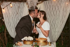 A couple share an intimate moment at their wedding dessert table featuring layered cakes under romantic string lights.