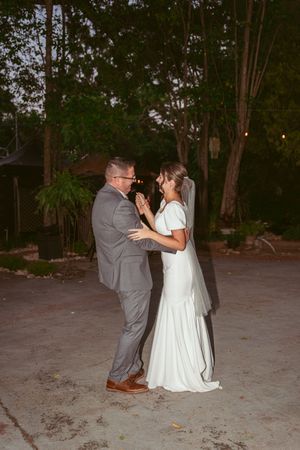 A romantic evening wedding dance between two people in formal attire under garden trees at dusk.