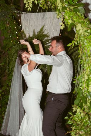 A couple in wedding attire dances playfully in a garden setting with string lights and greenery.