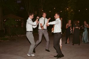 Three people in formal attire dancing energetically at an outdoor evening celebration while others watch from the background.