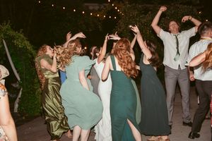 A lively group of wedding guests dance together at an outdoor evening reception under string lights.