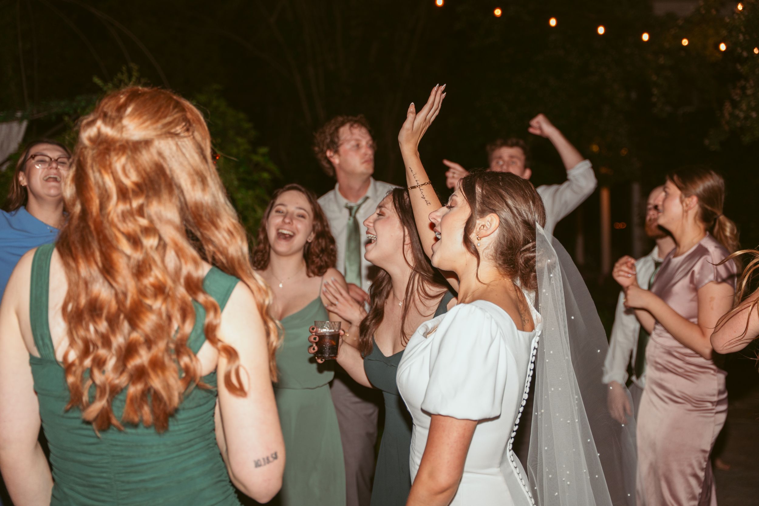 People dancing and celebrating at a lively wedding reception with twinkling lights overhead.