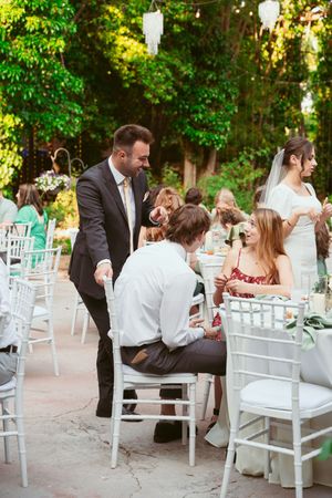 Wedding reception guests seated at elegant white tables with hanging lights in a garden setting.