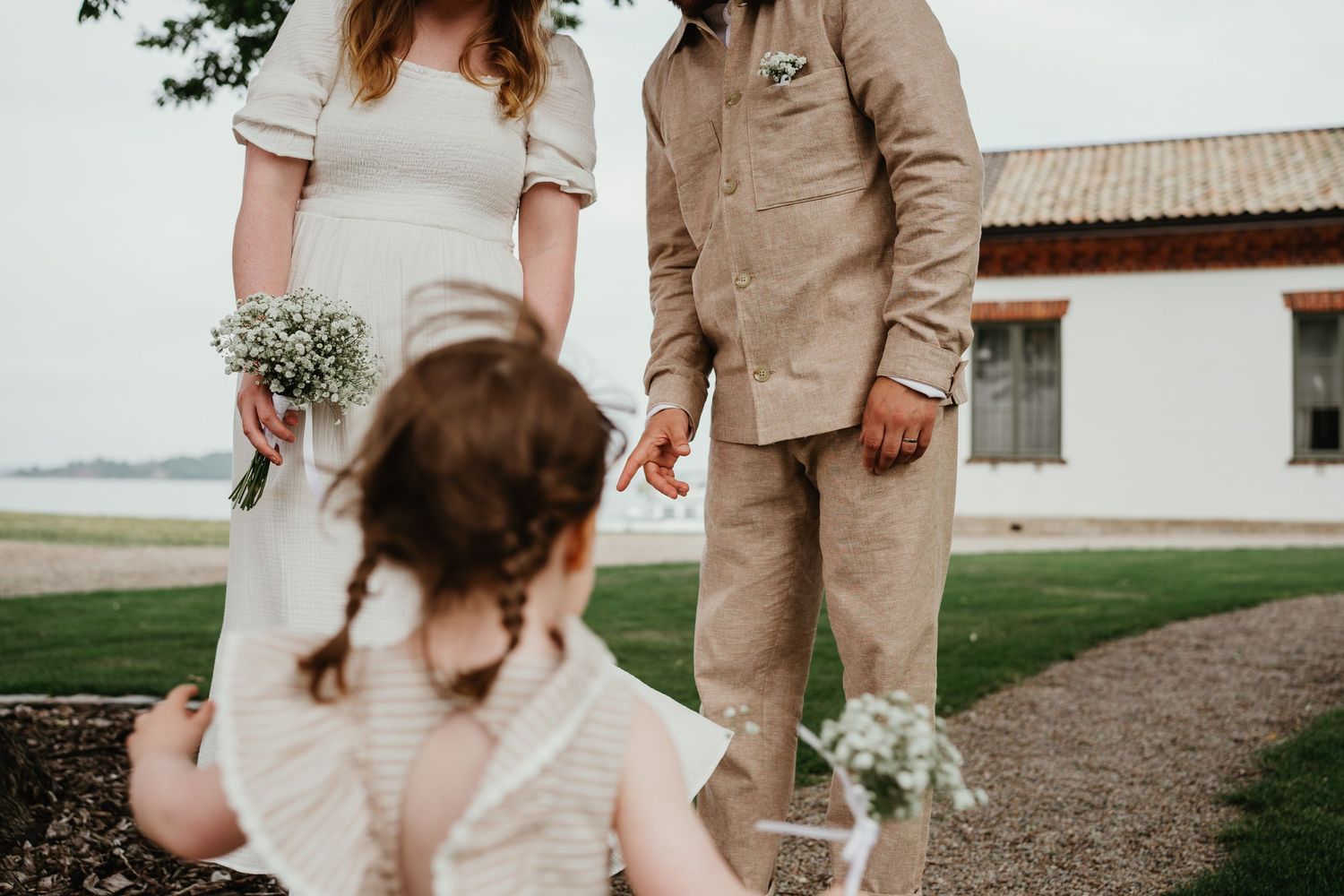 Small wedding ceremony takes place outdoors by the water on grassy lawn near white building.