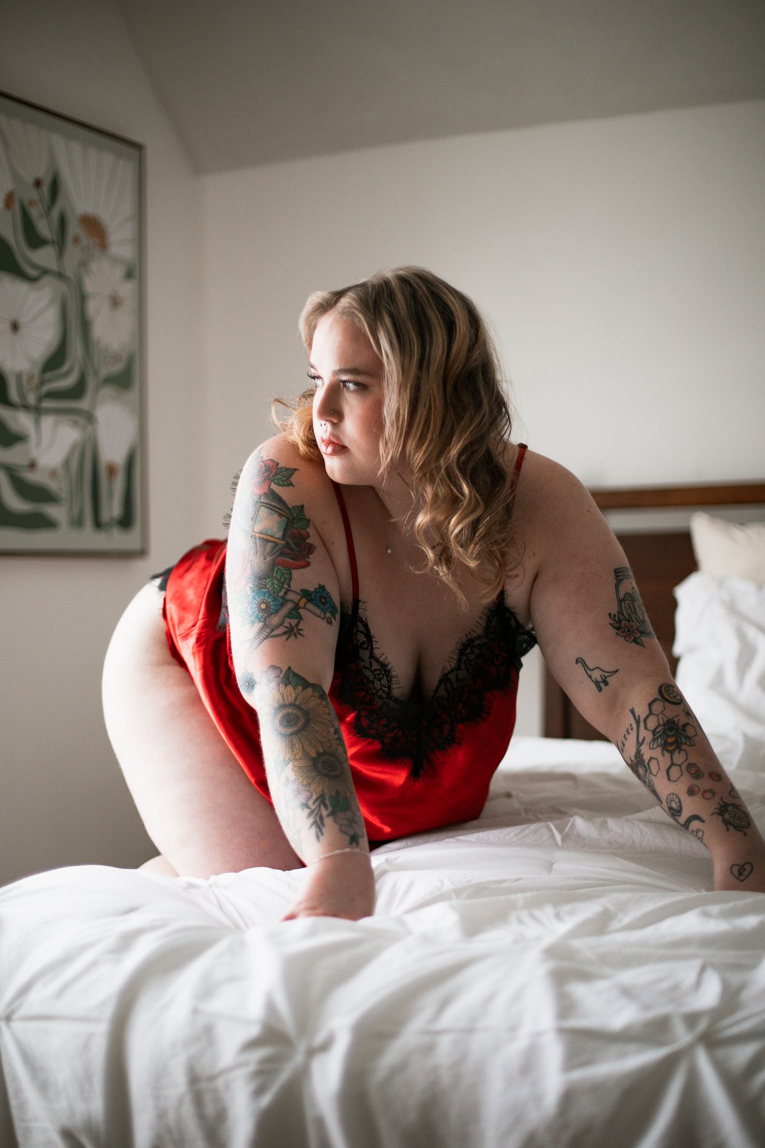 Artistic boudoir photography features someone in red lingerie with decorative arm tattoos posing on white bedding.