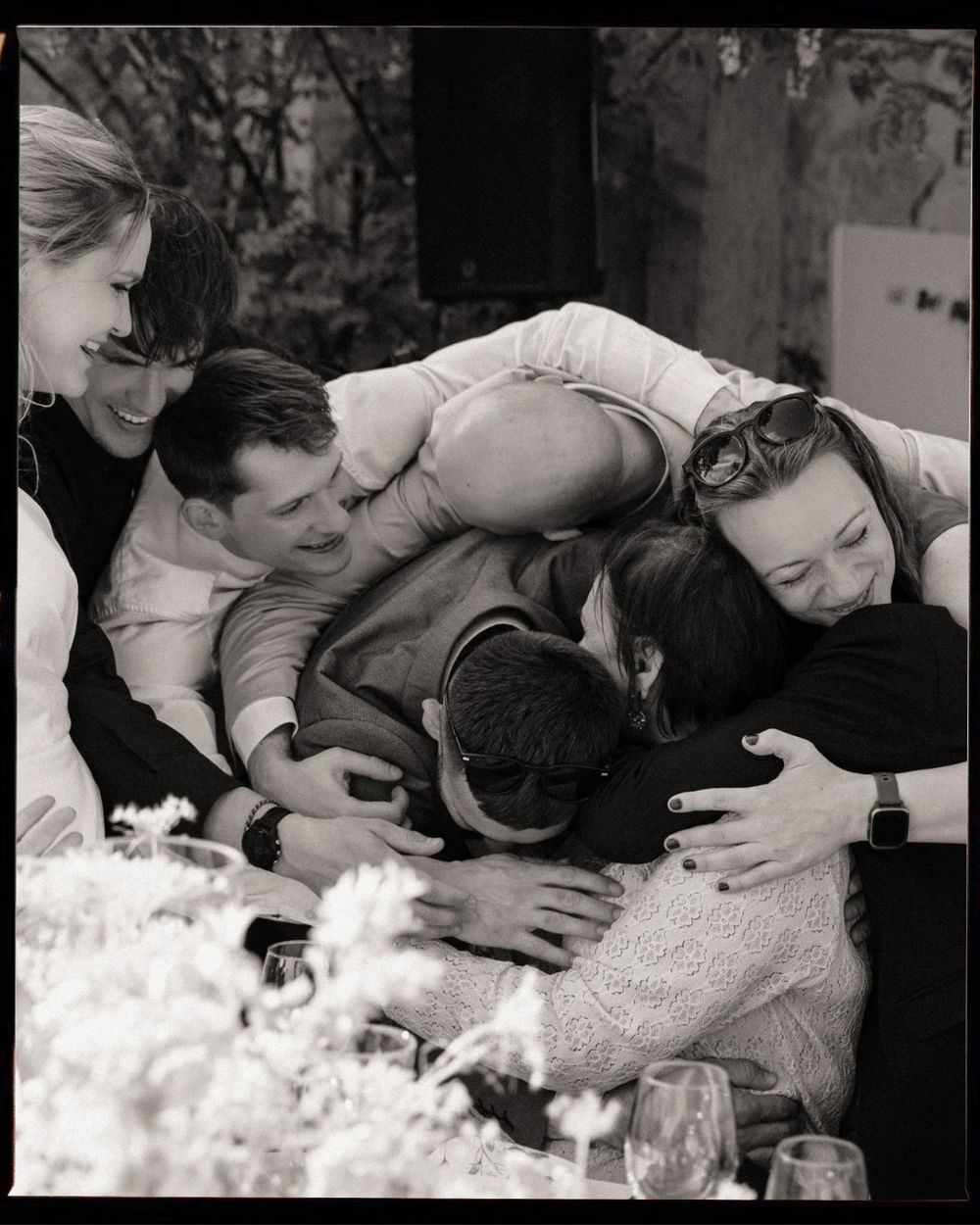 Black and white photo of a joyful group hug among friends at an outdoor wedding reception table.