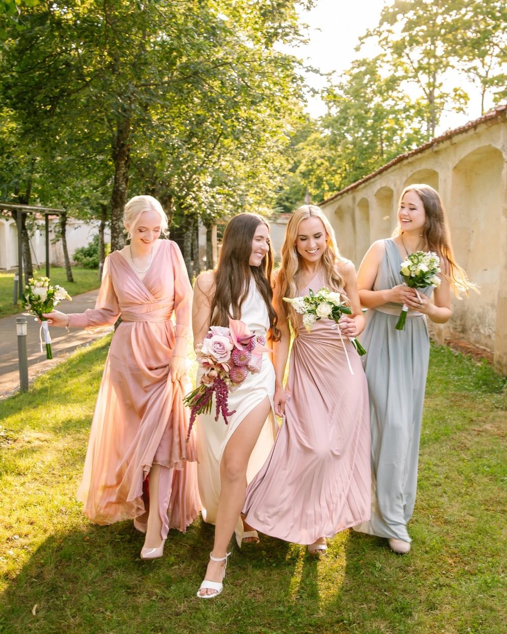 Four women in elegant gowns walk outdoors, with a bride in white and bridesmaids in pink and blue holding bouquets.