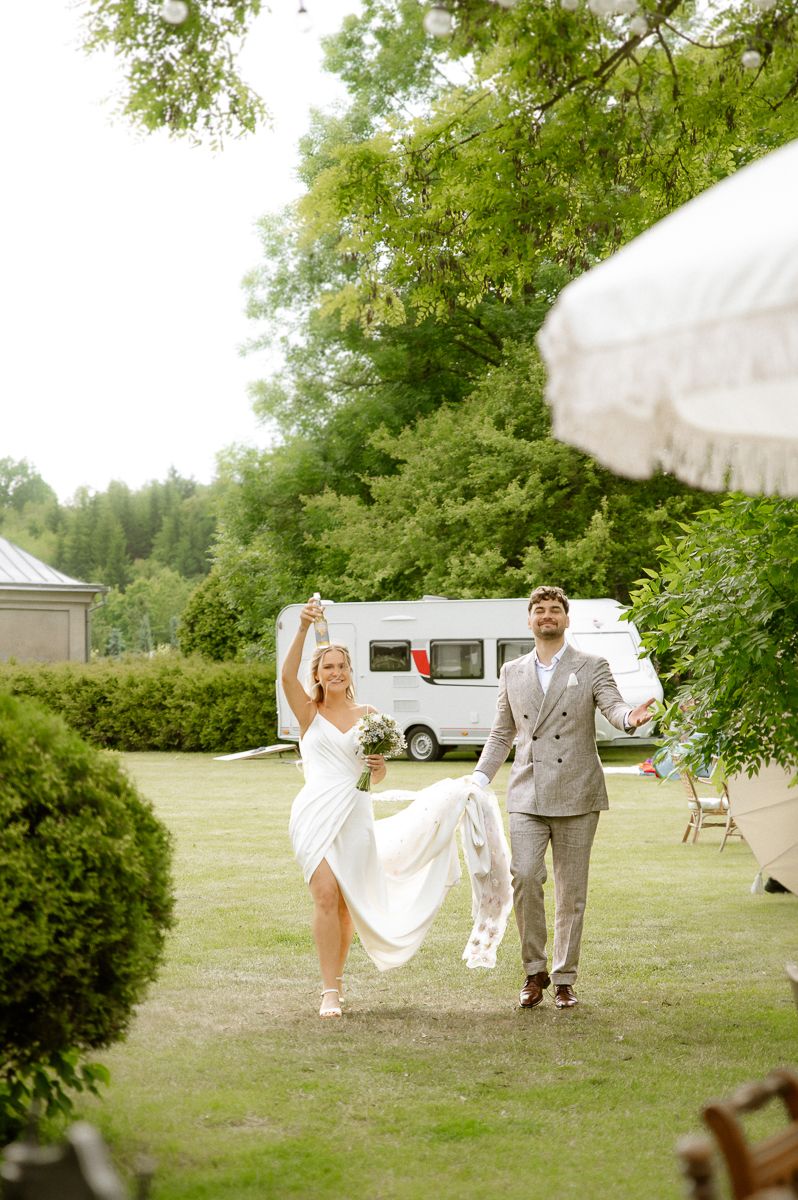 Joyful bride in white dress and groom in gray suit walking outdoors at garden wedding venue with trees and camper van.