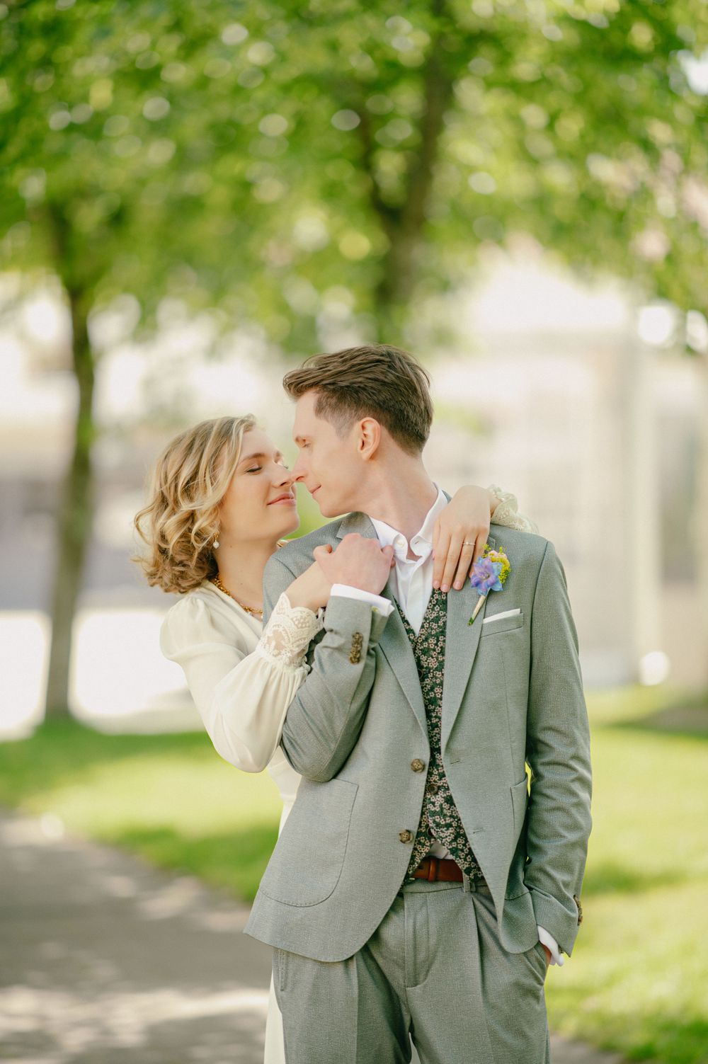 A romantic couple in gray attire share tender moments in a sunlit park with green trees in the background.