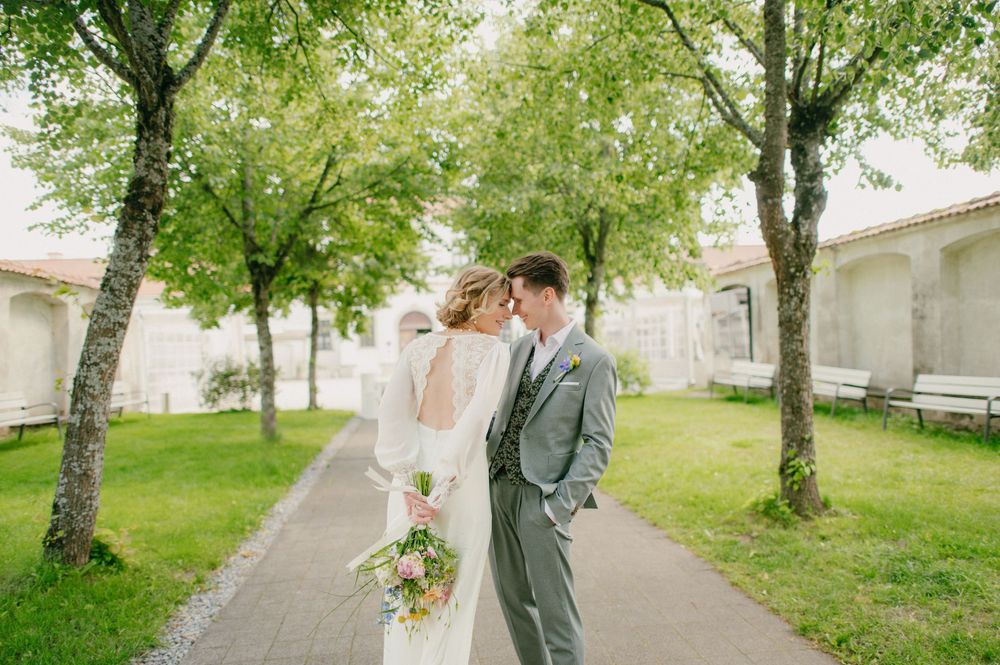 A couple shares a romantic moment in a lush garden setting with white buildings in the background.