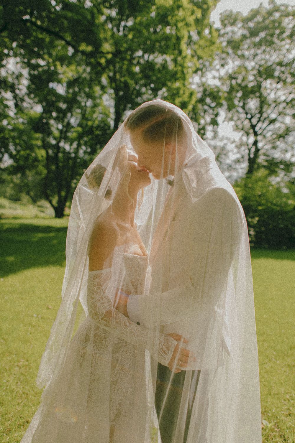 A flowing white veil billows in the breeze against a backdrop of sunlit trees in a park setting.