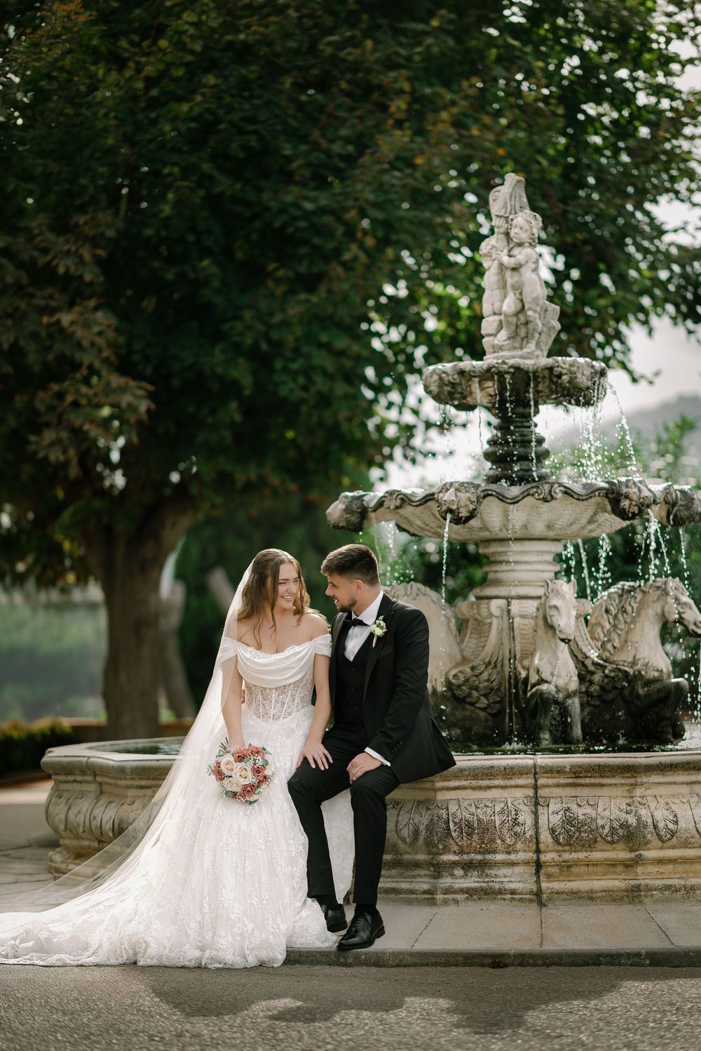 Bride and groom sitting together by an ornate stone fountain surrounded by lush green trees on their wedding day.