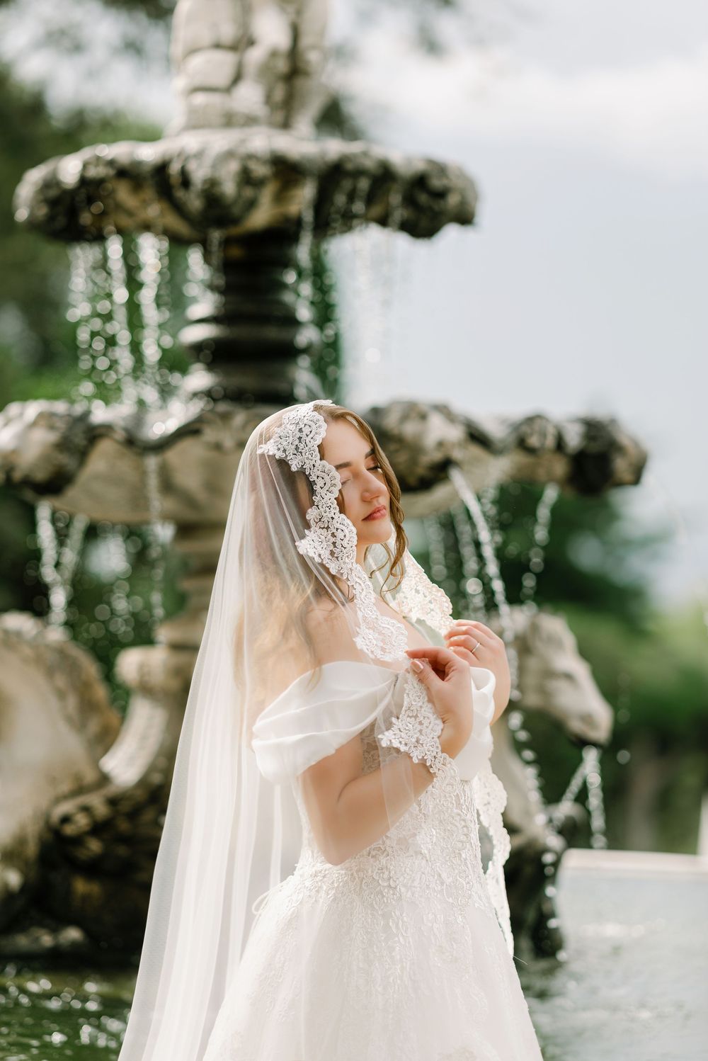 Elegant bride in lace veil and ball gown posing before ornate stone fountain in garden setting.