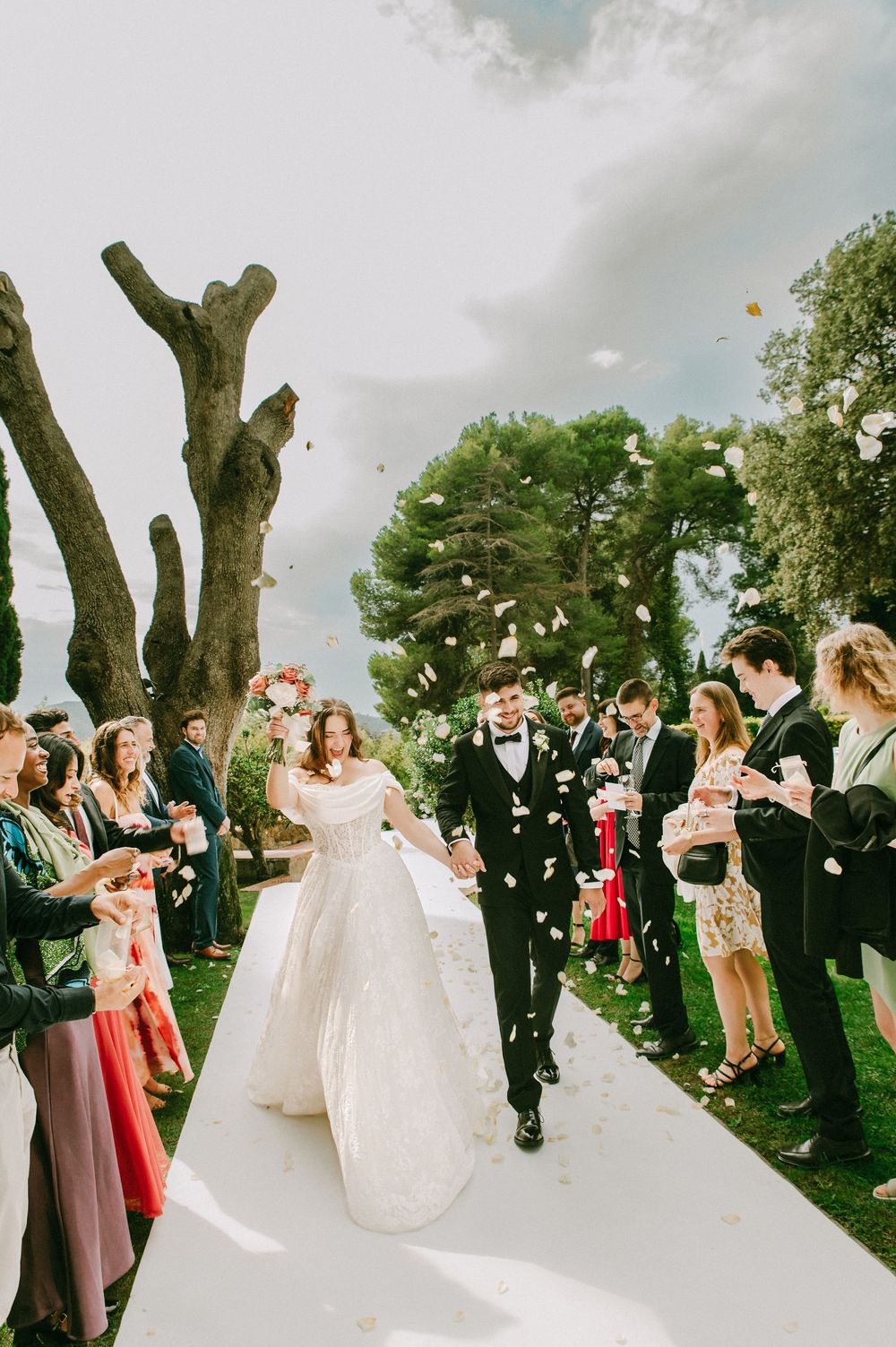 Bride and groom walk down outdoor aisle as guests toss flower petals at elegant garden wedding ceremony.