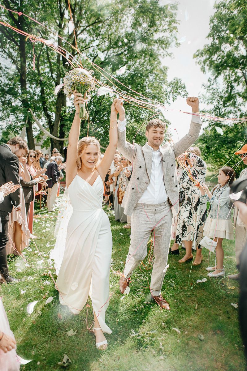 Wedding guests celebrate outdoors under green trees raising their hands joyfully in summer attire.