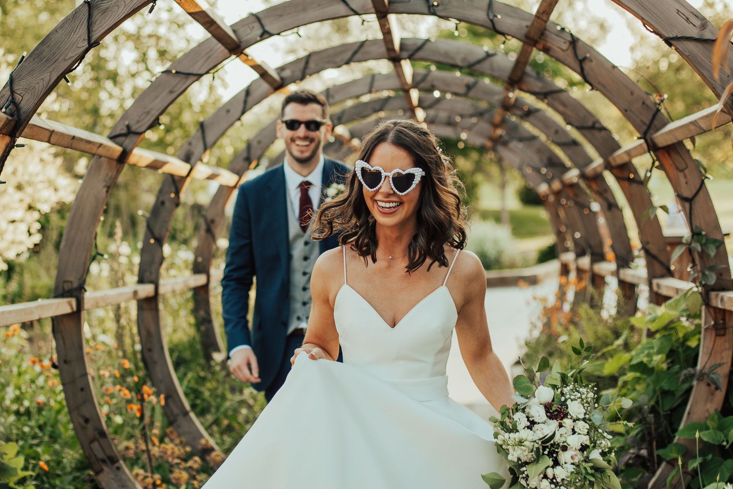 Newlywed couple walking hand in hand through scenic countryside field in formal wedding attire.