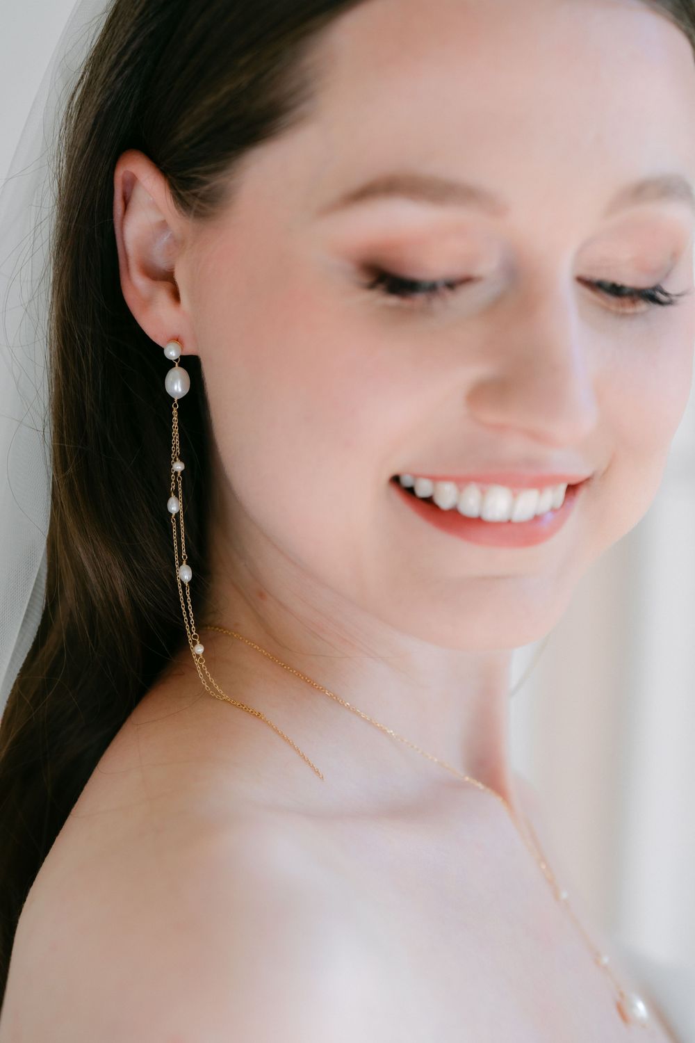 A close-up portrait showing a gentle smile and delicate pearl drop earrings against a soft, light background.