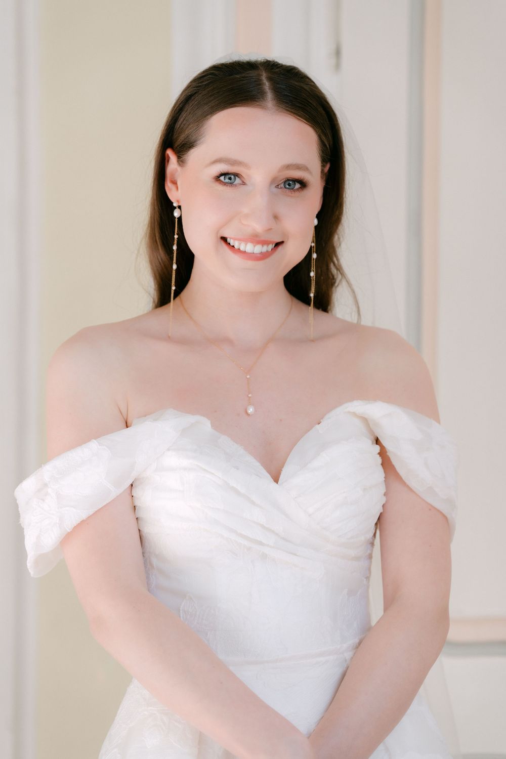 Bride in an elegant off-shoulder white wedding dress with delicate jewelry and natural makeup poses gracefully.