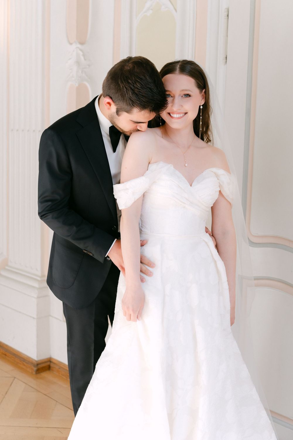 A bride in an off-shoulder white gown and groom in classic black tuxedo share an intimate moment in an elegant setting.