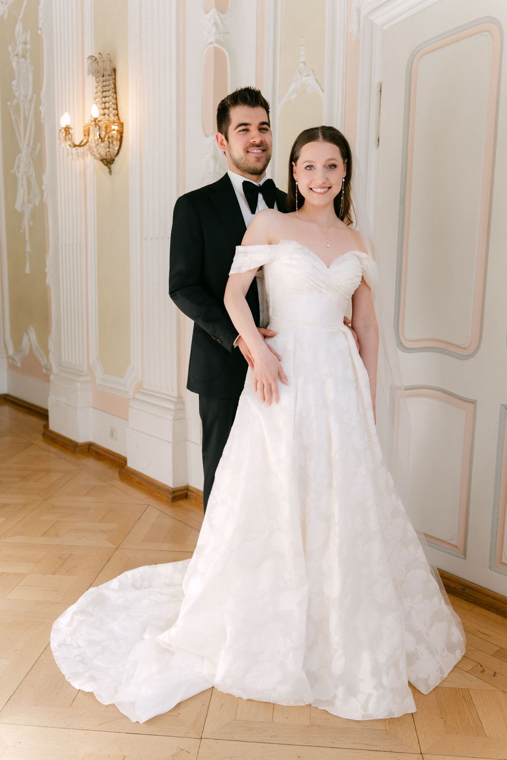 A bride in an off-shoulder white wedding gown stands with her groom in a black tuxedo in an elegant room with wall sconces.