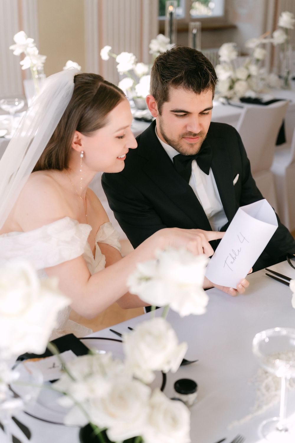 A couple at their wedding reception sit at an elegant white-decorated table reading a paper together.