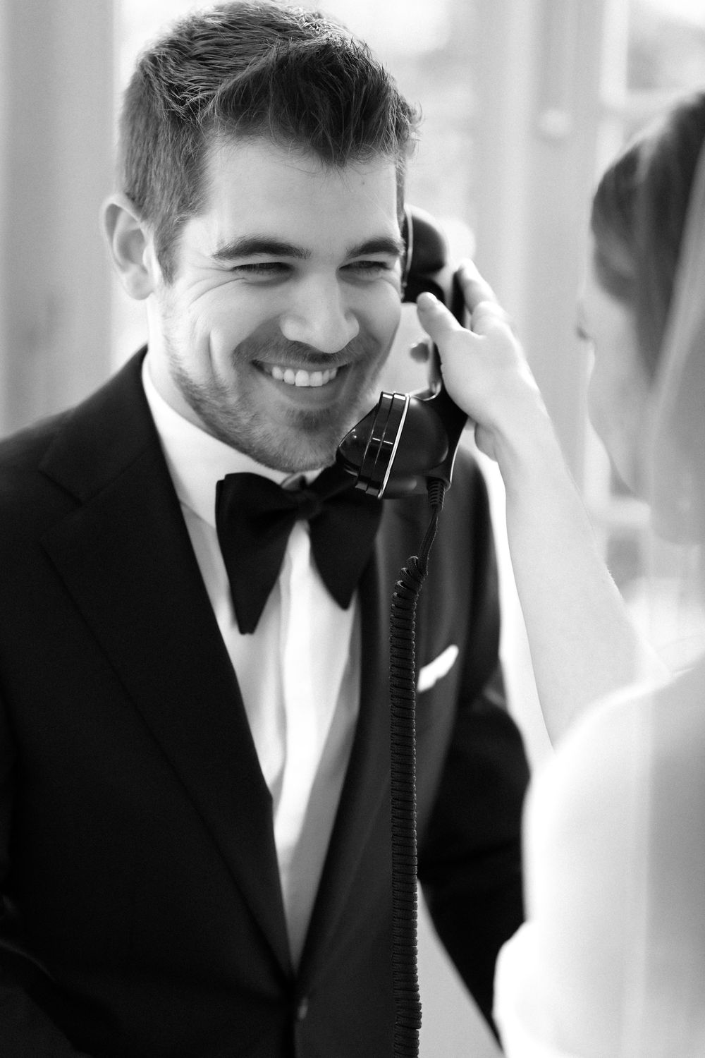 A person in a black tuxedo and bowtie smiles warmly while making a phone call at a formal event.