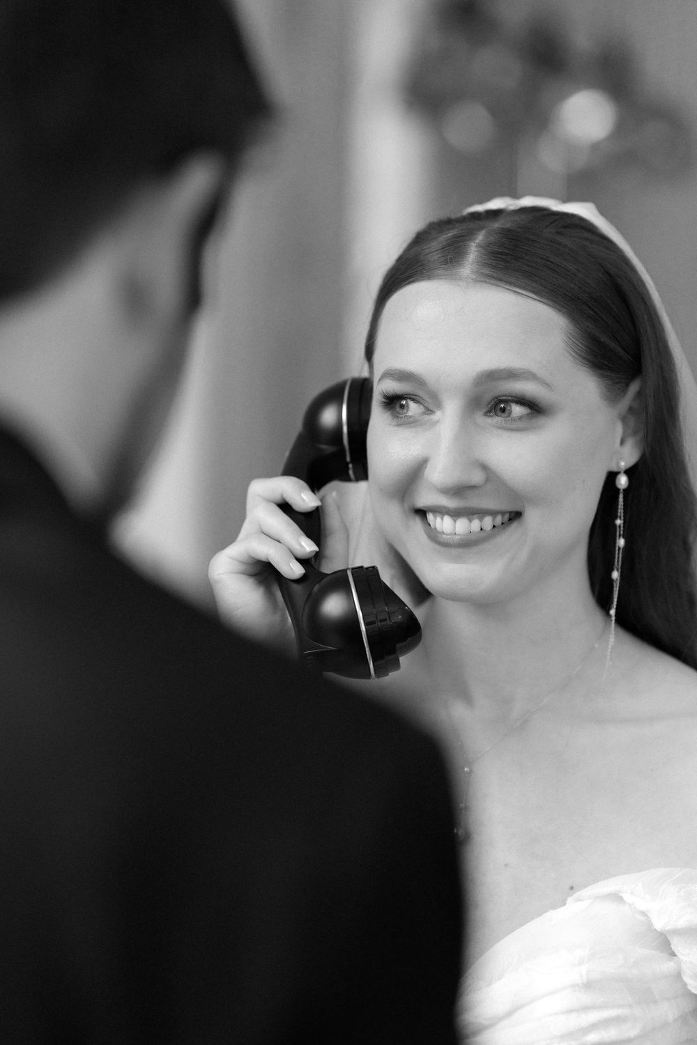 A bride in a white dress smiles warmly while speaking on a vintage black telephone during her wedding day.