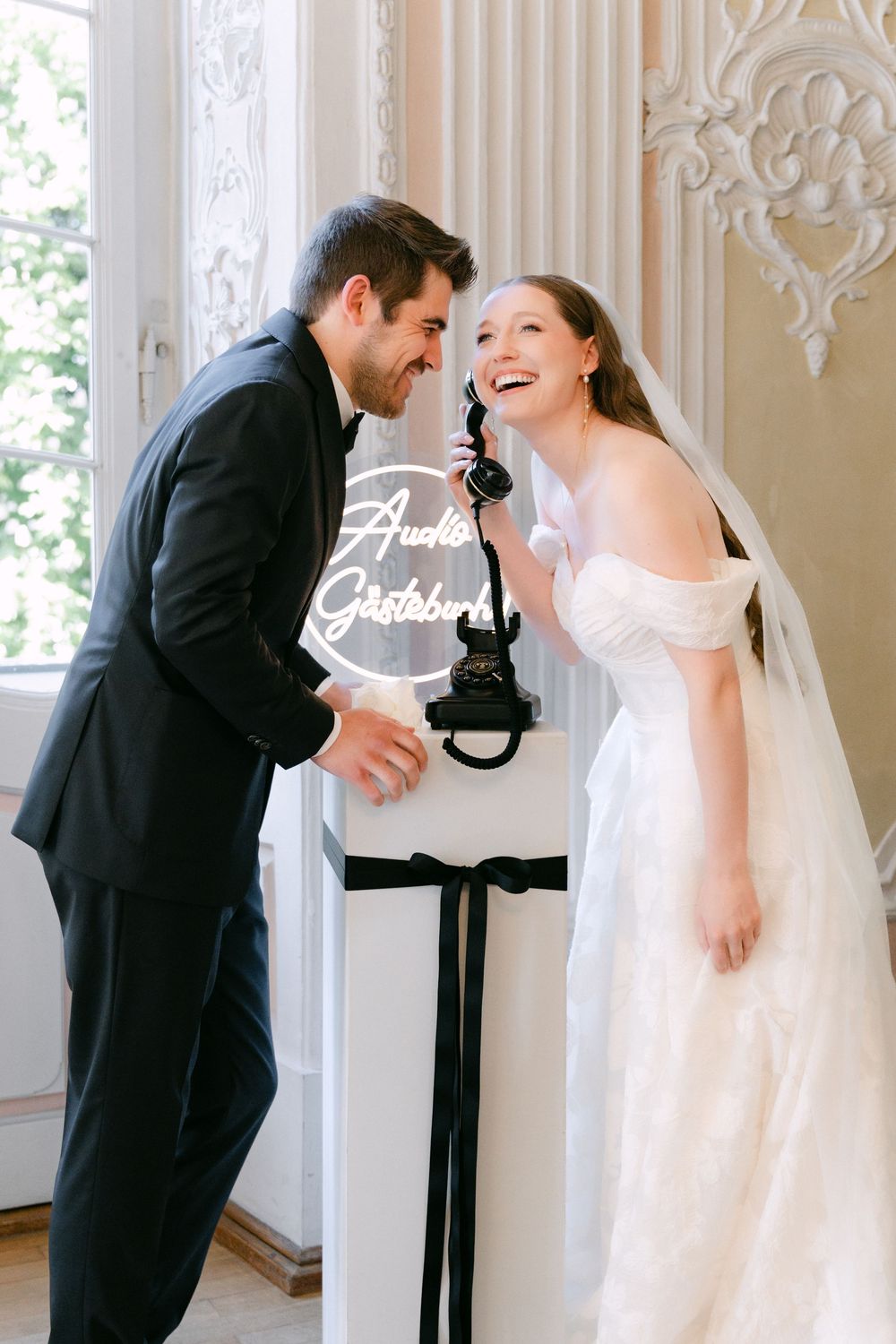 Happy couple in wedding attire laugh together beside an ornate decorative display stand in an elegant white room.