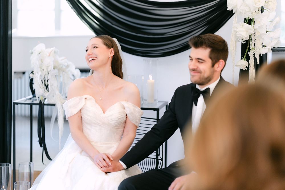 A couple sits together in their wedding attire, with elegant white floral arrangements and black drapes in the background.
