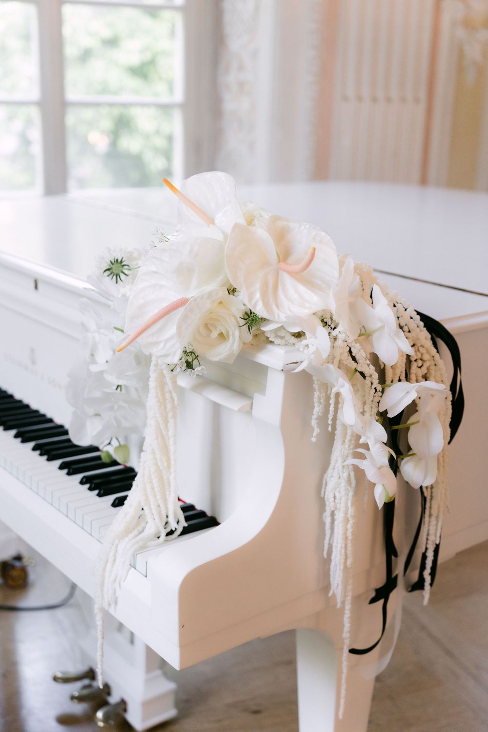 Elegant white floral arrangement with cascading blooms decorates the corner of a white piano in a bright sunlit room.