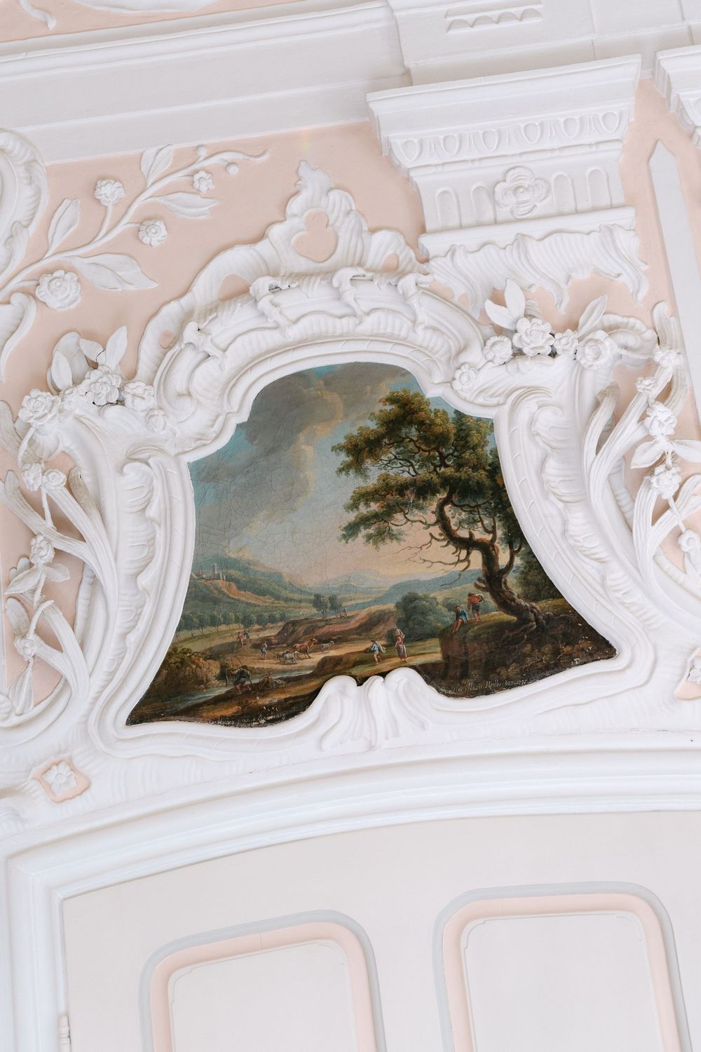 Ornate white rococo plasterwork frame surrounds a pastoral landscape painting on a pale pink wall.