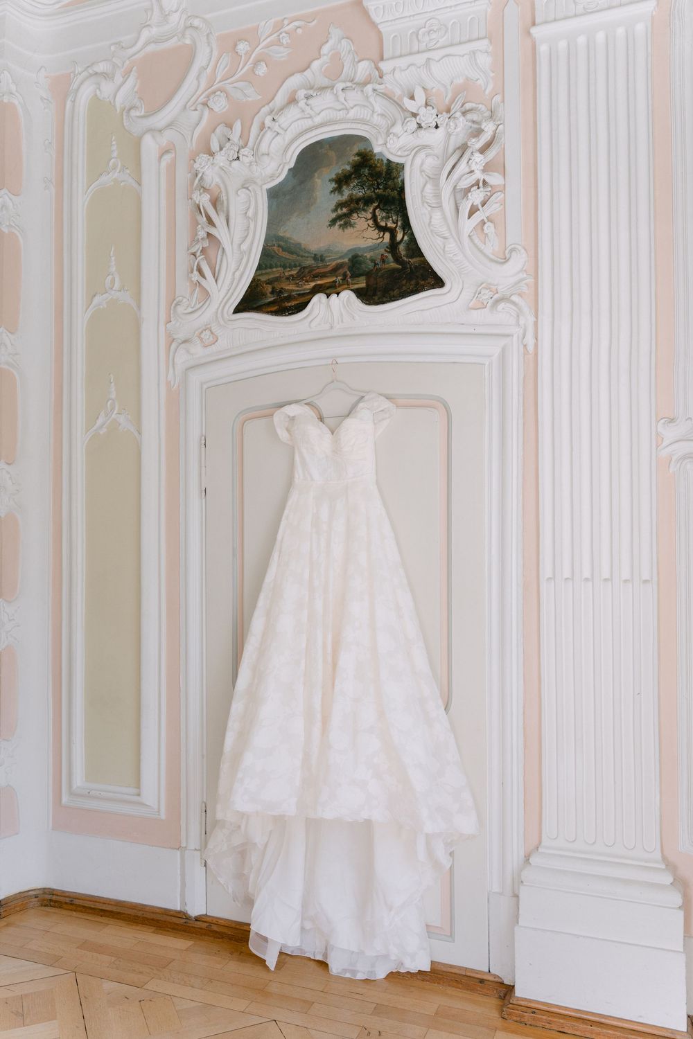 An elegant white lace wedding dress hangs on an ornate wall beneath a framed landscape painting in a classical setting.