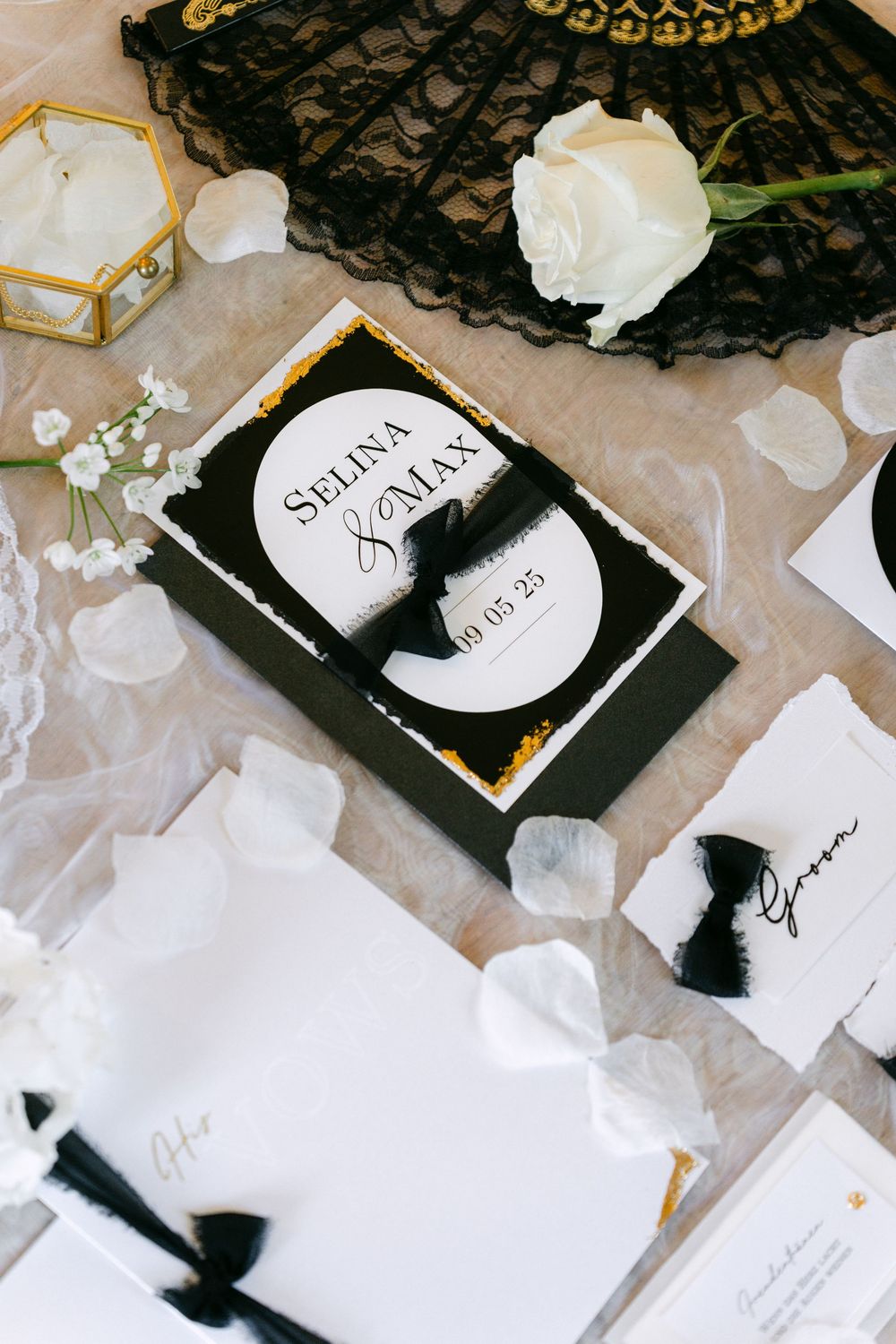Black and white wedding invitation with elegant gold accents, white rose, and lace details on textured paper.