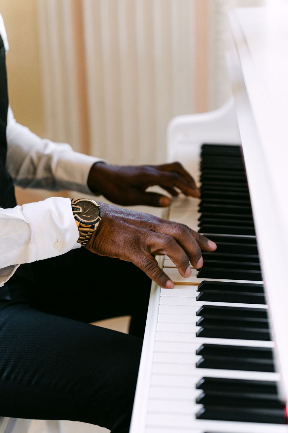 Hands wearing gold watch play piano keys on white keyboard in elegant indoor setting.