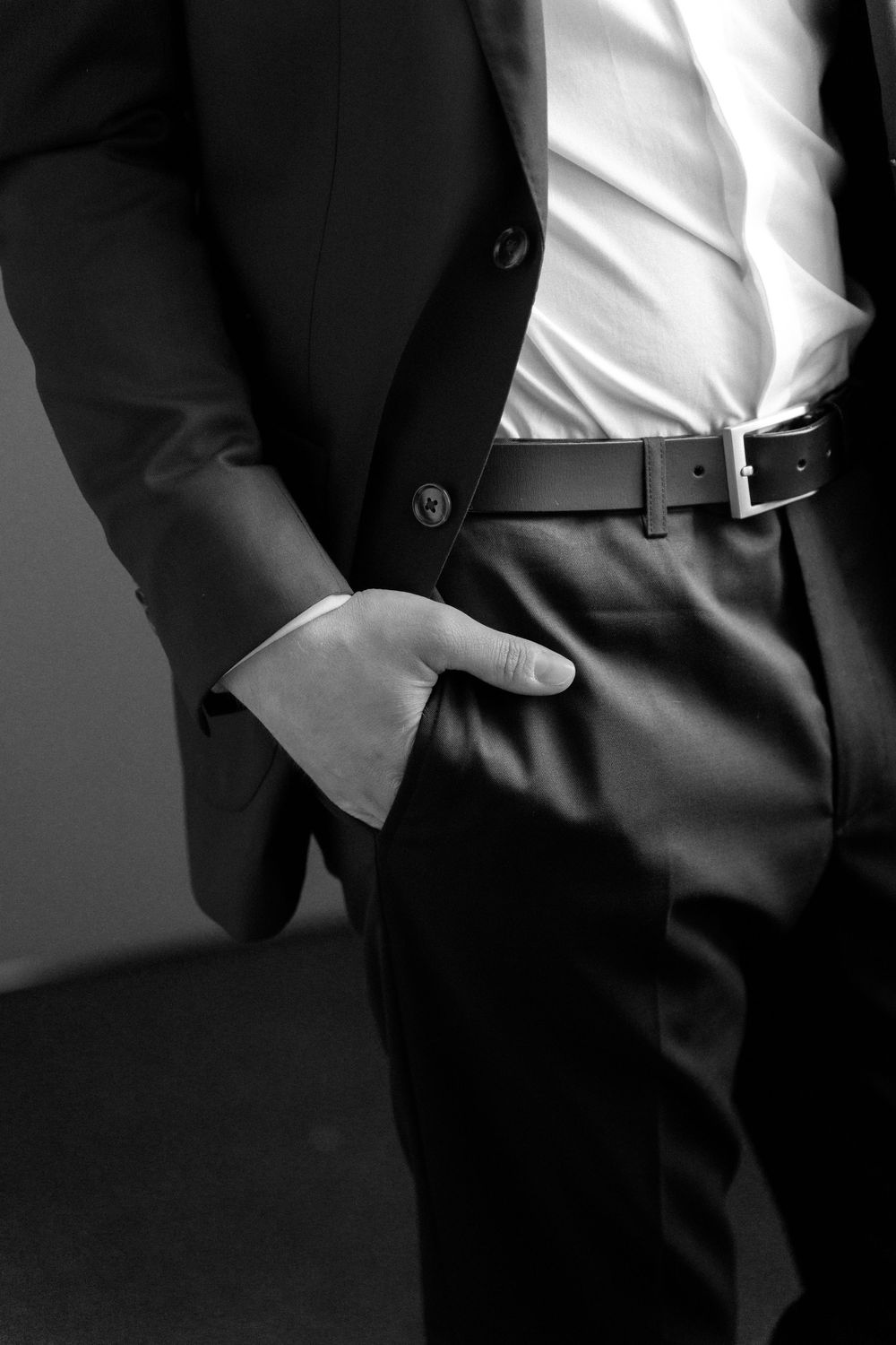 Black and white fashion detail shot of a man in formal attire with blazer, dress shirt and belt against dark background.