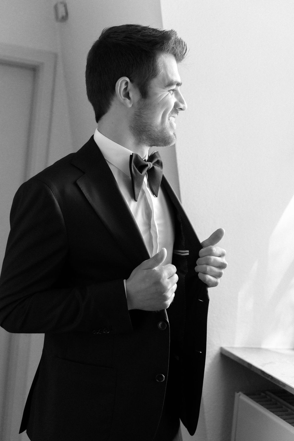 Black and white profile shot of someone wearing a classic tuxedo with bow tie, adjusting their jacket while smiling.