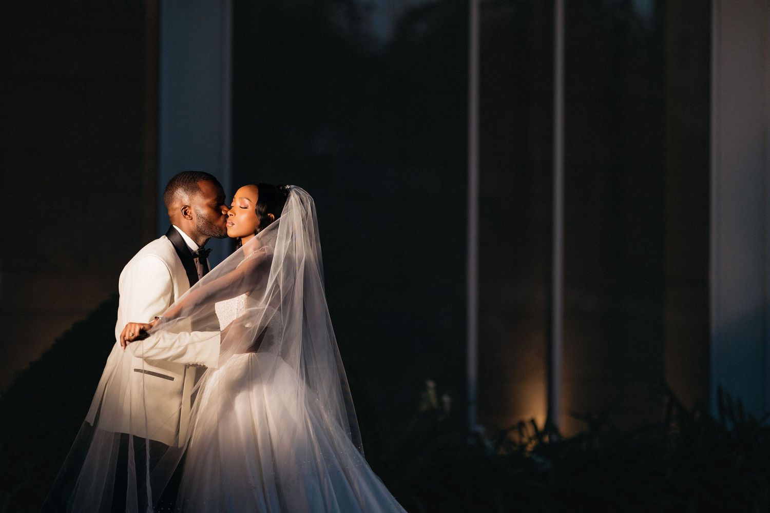 A newlywed couple shares an intimate moment in dim lighting, with the bride wearing a flowing white veil.