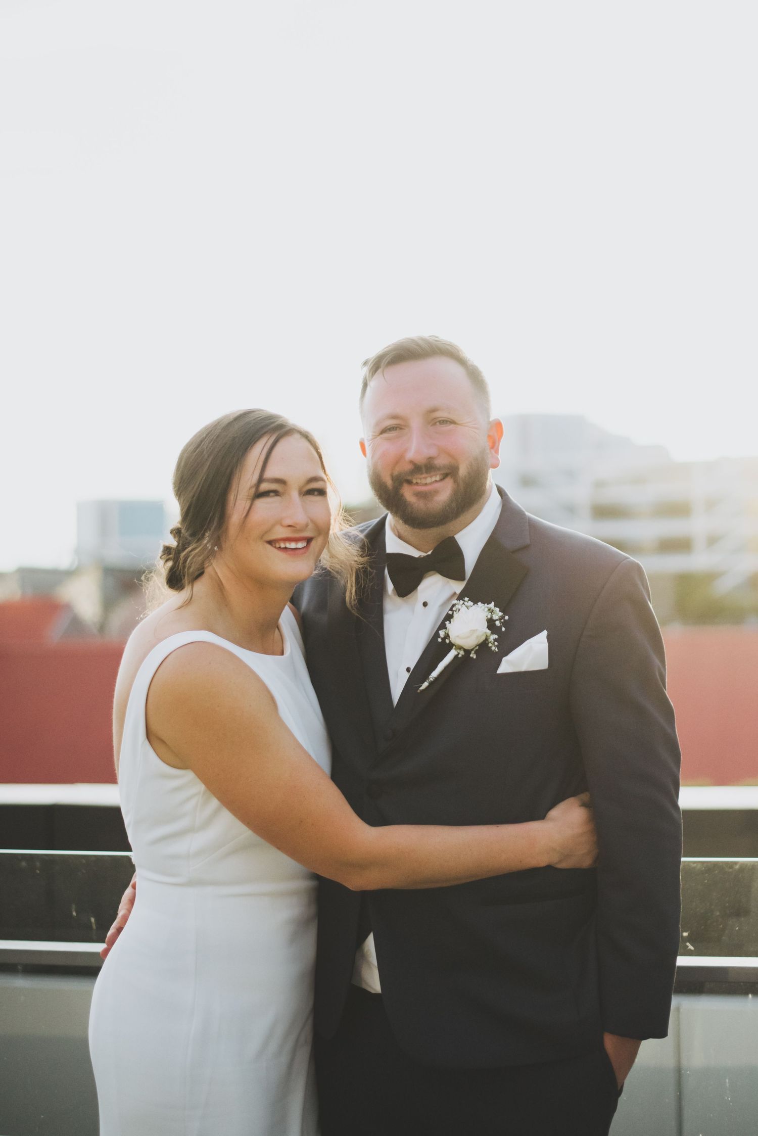 A happy couple poses together in formal wedding attire on a rooftop during sunset with city buildings in the background.