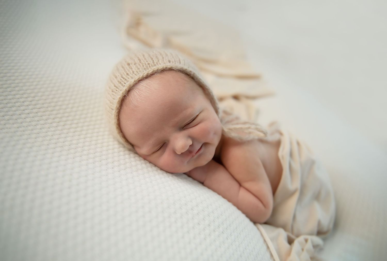 Sleeping newborn baby wrapped in cream colored fabric resting on textured white surface.