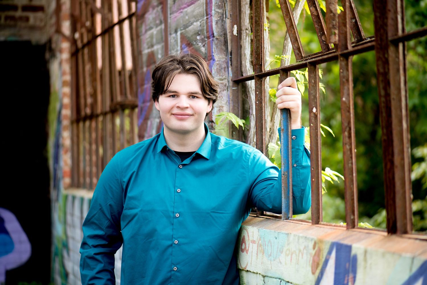 Person wearing a teal button-down shirt poses against a rustic backdrop with graffiti.