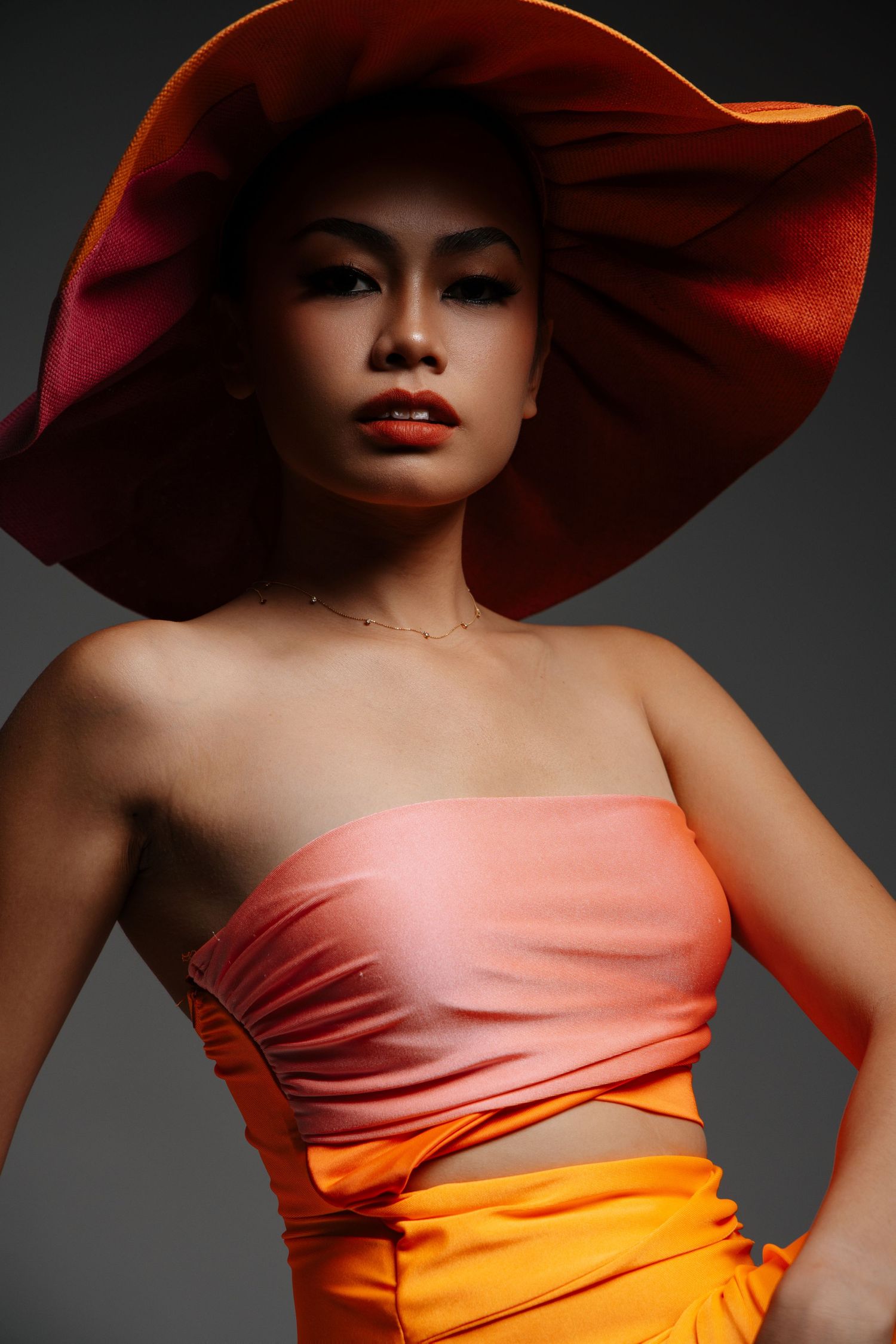Fashion portrait with dramatic red hat and orange dress against dark background.
