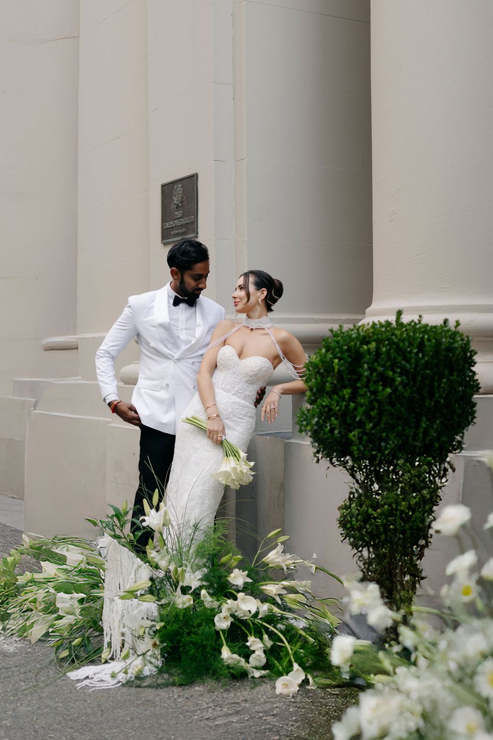 Wedding couple poses by columned building with white floral arrangements.