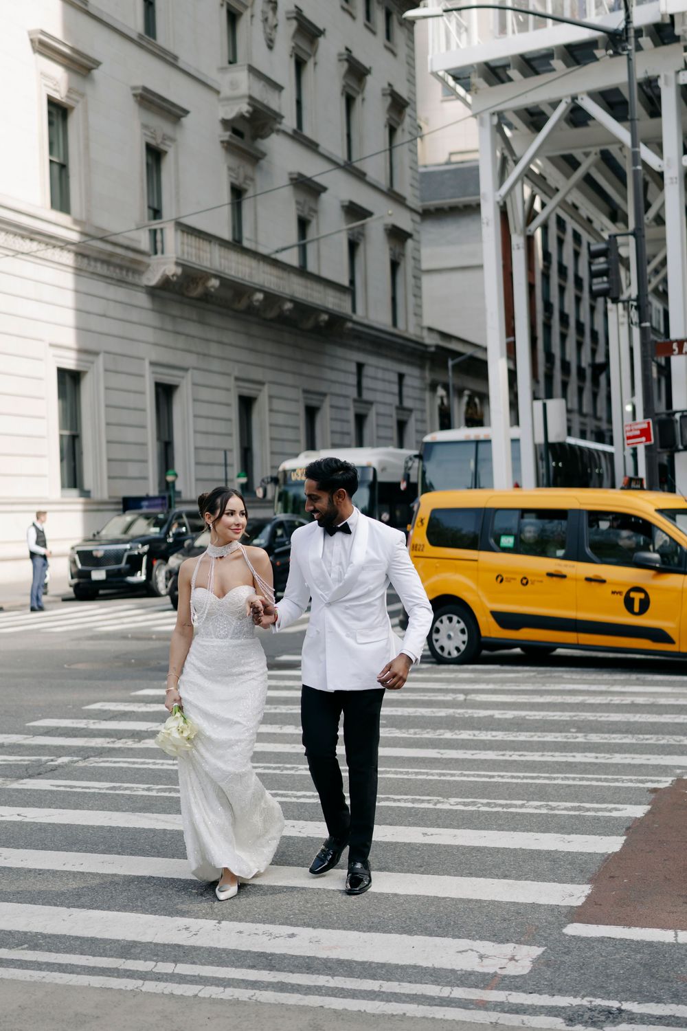 Couple walking across a city crosswalk with yellow taxi in background during wedding photoshoot.