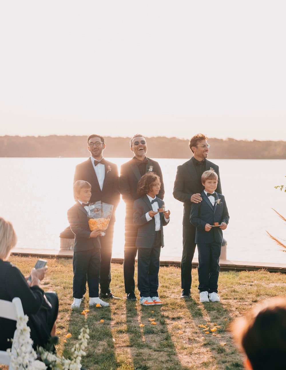 Wedding party members in formal attire standing on grassy lawn at sunset by the water.