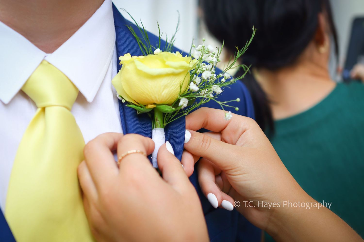 Hands pin a yellow rose boutonniere on a navy suit jacket with matching yellow tie at a formal event.